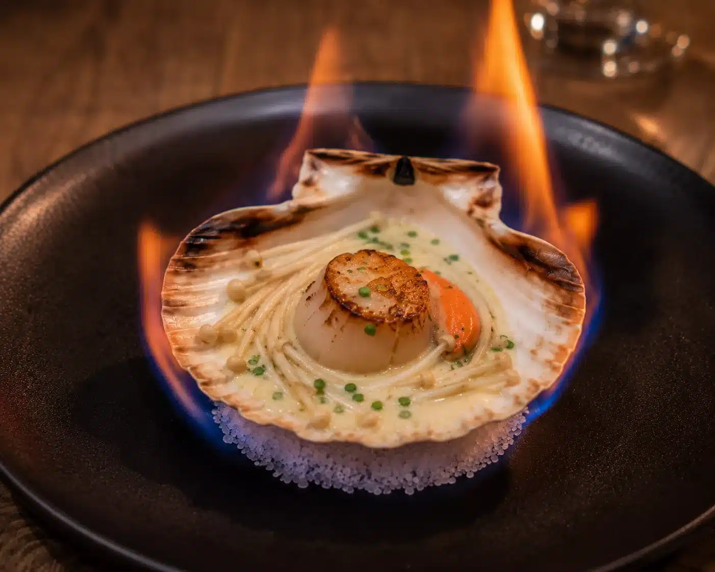 Jacobsmuscheln zuerst gegrillt, dann mit Sahne Champagner Sauce und Enoki Pilzen verfeiert. Wir servieren auf mit Brennalkohol benässtes grobes Meersalzbett und entzünden es im Moment des Servierens